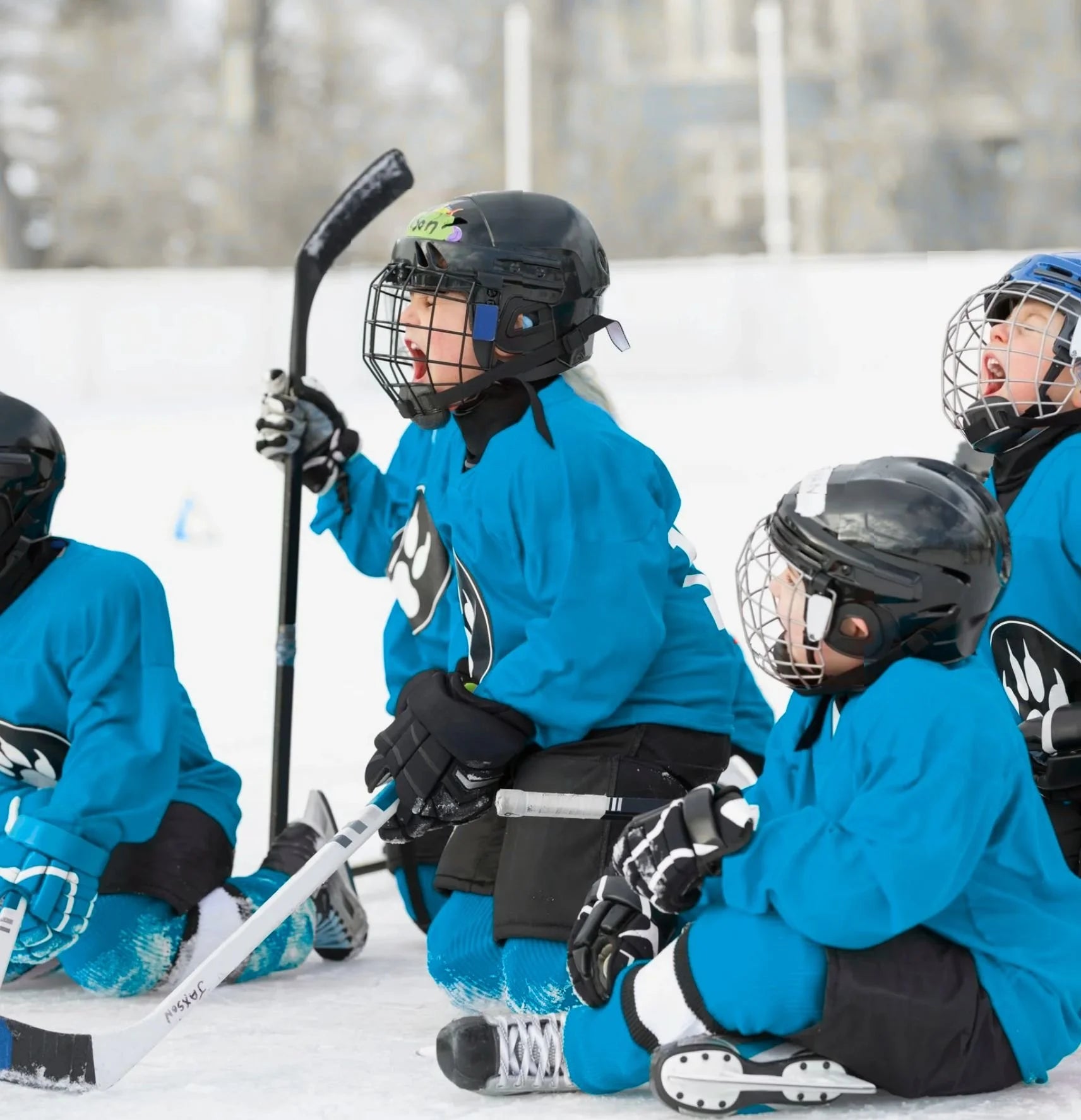 Children in blue hockey jerseys with black helmets and skates sitting on a snowy surface.